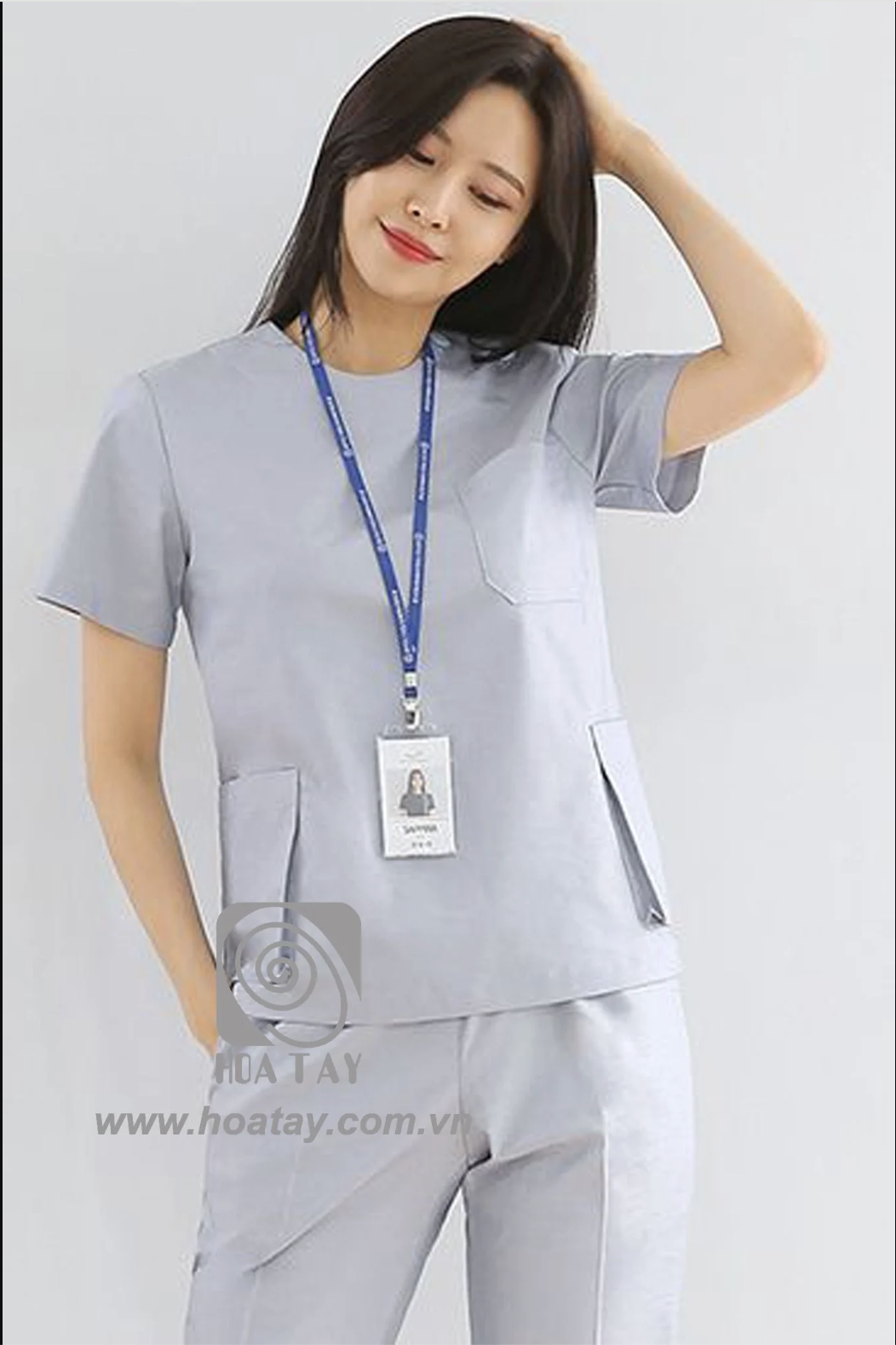 Một nữ nhân viên y tế đang đứng, mặc bộ đồng phục màu xám nhạt (light grey), áo cổ tròn tay ngắn với túi ngực và hai túi hông lớn, chất liệu vải trơn, đeo thẻ tên (ID badge) có dây đeo màu xanh, phông