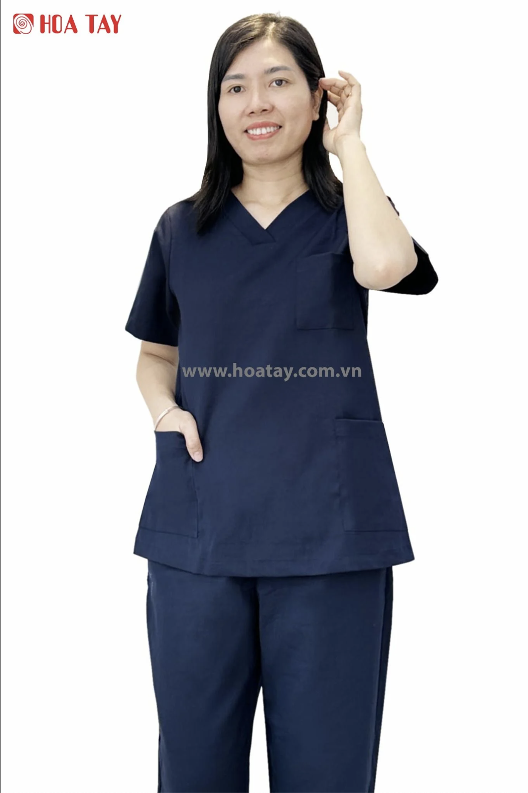 Một nữ nhân viên y tế đang đứng, mặc bộ đồ scrubs (đồng phục phẫu thuật) màu xanh navy, cổ chữ V, tay ngắn, có túi ngực và túi hông, chất liệu vải cotton pha, nền trắng sáng.