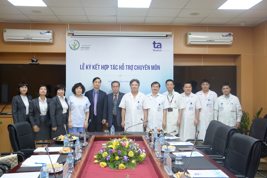 Nhóm 12 người đứng trước backdrop in chữ 'Lễ ký kết hợp tác hỗ trợ chuyên môn' và logo 'Việt Đức' cùng 'Tâm Anh'. Ba người mặc vest, một người mặc áo thun trắng, tám người mặc áo blouse trắng. Họ đứng