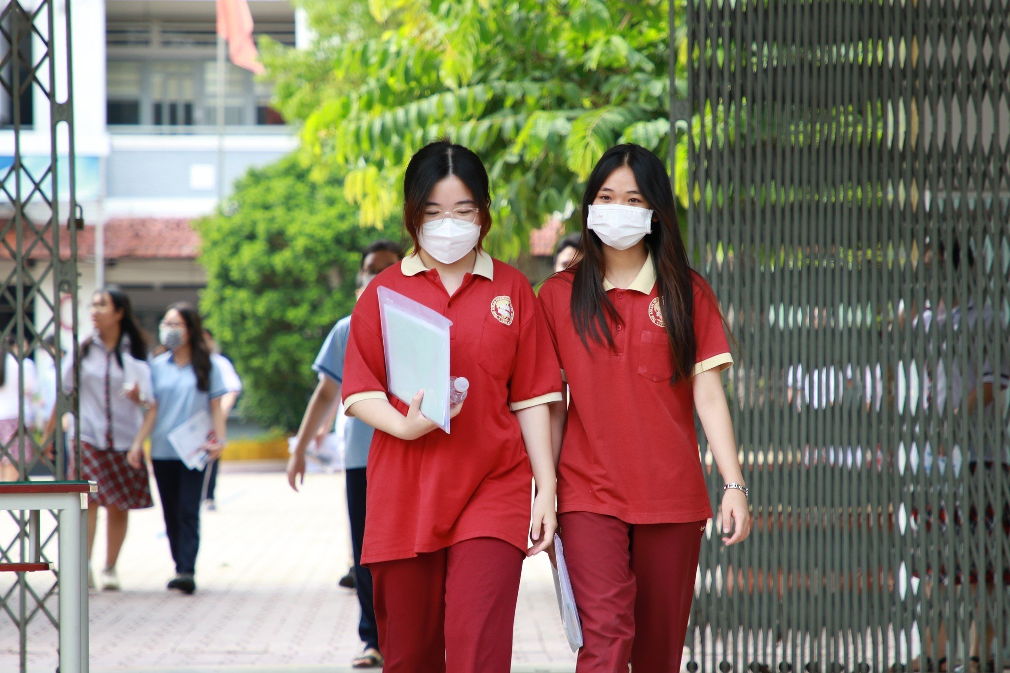 Hai nữ sinh đang đi bộ, mặc áo polo (áo cổ bẻ) màu đỏ phối cổ vàng, quần dài cùng tông, đeo khẩu trang y tế trắng, cầm tài liệu, bối cảnh sân trường ngoài trời dưới ánh sáng tự nhiên.