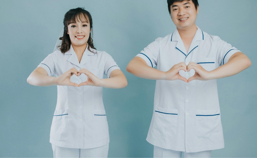 Hai nhân viên y tế (một nam, một nữ) tạo hình trái tim bằng tay trước ngực. Họ mặc bộ đồng phục y tế màu trắng, cổ bẻ, tay ngắn, có đường viền xanh dương ở cổ, tay áo và túi. Nền màu xanh xám, ánh sán