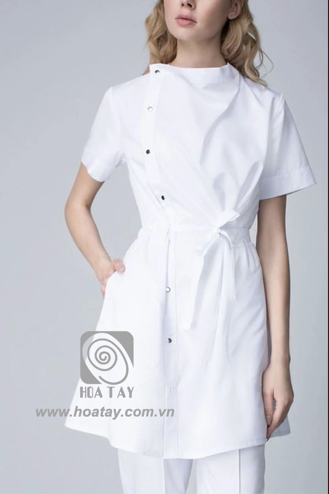 Một nữ nhân viên y tế mặc áo blouse trắng (áo bác sĩ) tay ngắn, cổ chéo (cổ tàu lệch), cài khuy bấm (nút bấm kim loại), có dây thắt eo (đai buộc) và túi hông, chất liệu vải cotton phẳng, phông nền xám