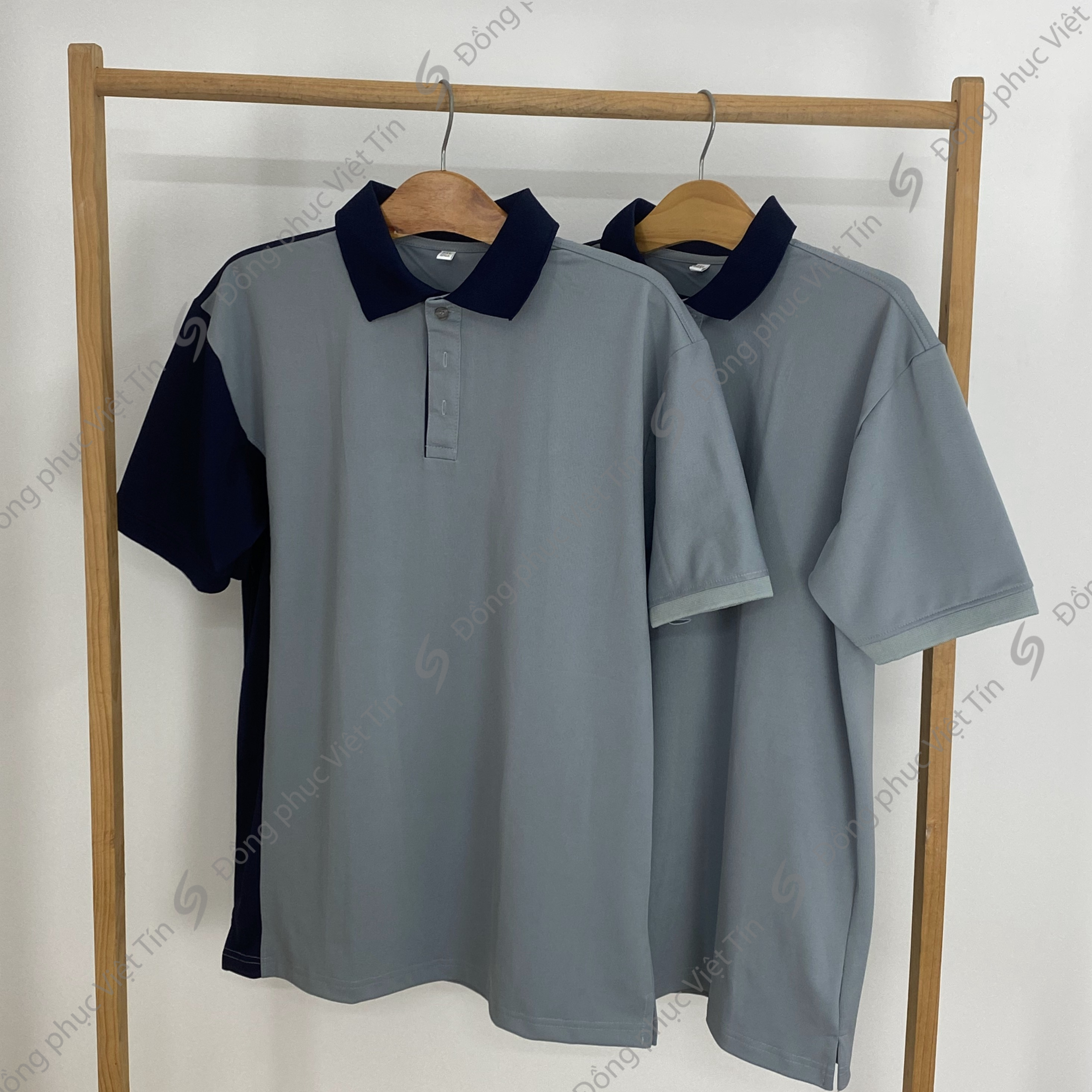 Hai chiếc áo polo phối màu xám và xanh đen chất liệu Poly tiêu chuẩn, treo trên móc gỗ, khung treo bằng gỗ trên nền trắng, bố cục tối giản, chuyên nghiệp.