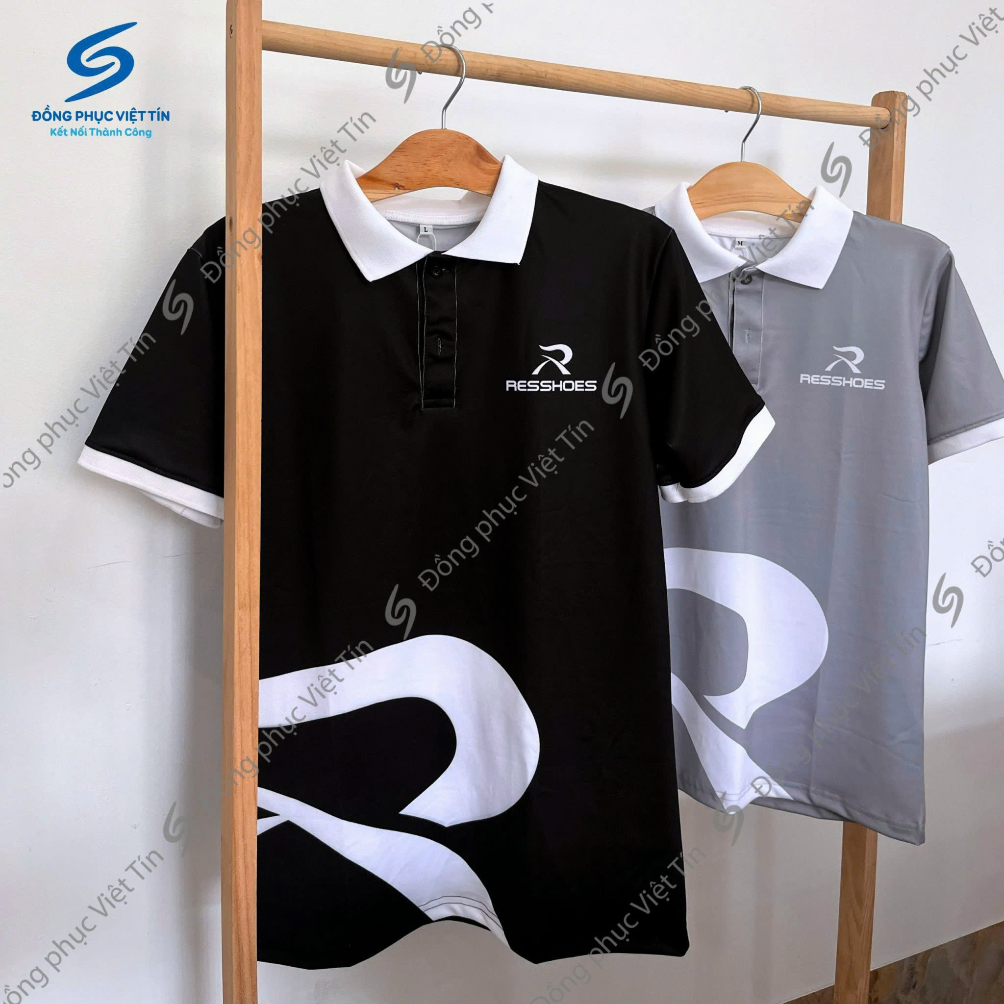 Hai chiếc áo polo đồng phục treo trên giá gỗ, một chiếc màu đen và một chiếc màu xám, chất liệu Poly tiêu chuẩn, được in 3D họa tiết trên áo và logo RESSHOES in nổi bật. Nền tường trắng sáng, bố cục tối giản, chuyên nghiệp.