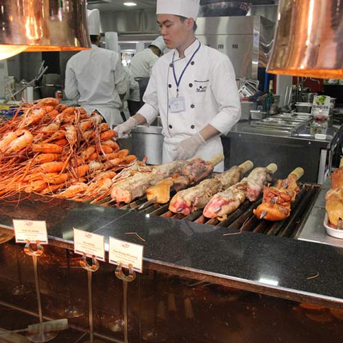 Một đầu bếp nam đang nướng thịt; mặc áo khoác trắng hai hàng khuy (double-breasted), găng tay dùng một lần, mũ toque vải, bảng tên đeo cổ; bối cảnh bếp nhà hàng chuyên nghiệp, ánh sáng vàng từ đèn hâm