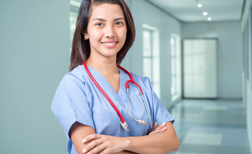 Một nữ y tá đứng hai tay bắt chéo trước ngực, mỉm cười nhìn thẳng ống kính. Cô mặc bộ scrubs y tá màu xanh dương nhạt, cổ chữ V, tay ngắn, chất liệu vải mềm. Quanh cổ là ống nghe màu đỏ đầu kim loại b