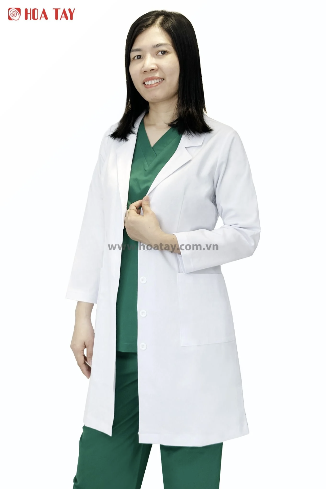 Một nữ nhân viên y tế đang đứng, mặc áo blouse trắng (áo khoác bác sĩ) dài tay, cổ bẻ (ve áo), cài khuy giữa, chất liệu vải phẳng phiu, bên trong là bộ đồ scrub màu xanh lá, nền trắng sáng.
