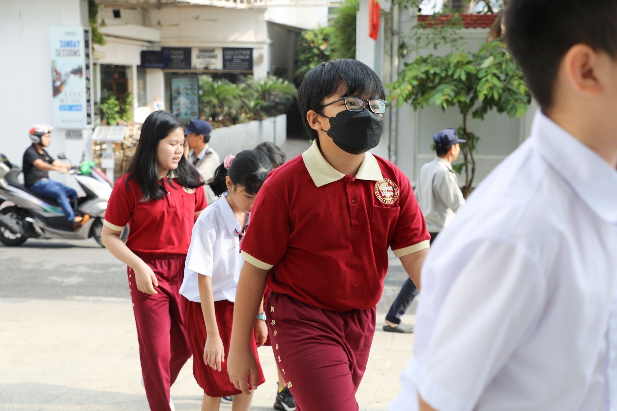 Một nhóm học sinh (4 em) đang đi bộ trên vỉa hè; mặc áo polo đỏ cổ kem (cổ bẻ) và quần dài màu đỏ đô (quần tây), đeo khẩu trang y tế đen, kính cận; bối cảnh đường phố ban ngày, ánh sáng tự nhiên.