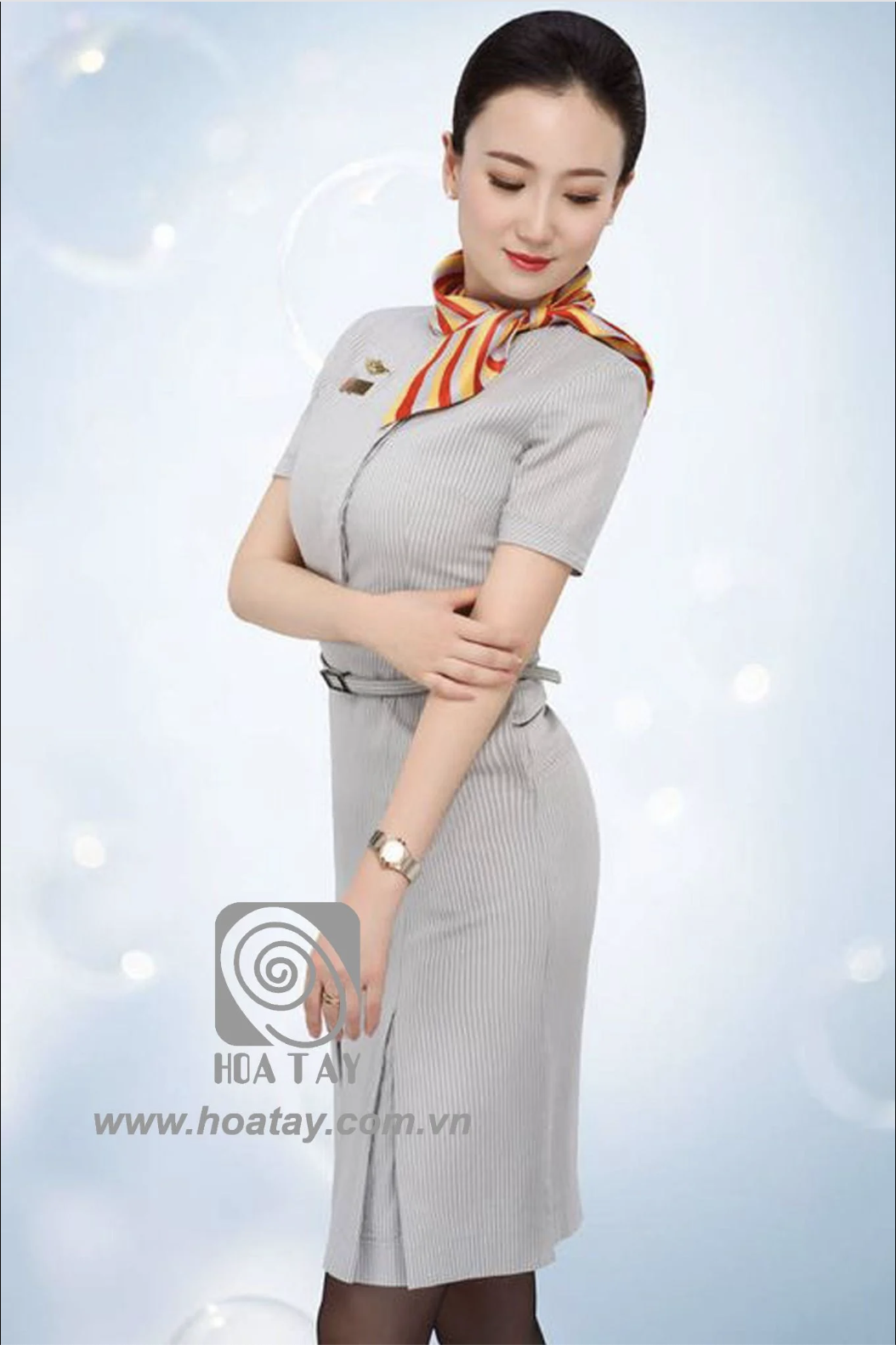 Một nữ tiếp viên hàng không đứng nghiêng, mặc váy liền thân (dress) màu xám kẻ sọc, thắt lưng mảnh, khăn quàng cổ đa sắc, huy hiệu ngực, xẻ tà trước, nền xanh nhạt ánh sáng studio.