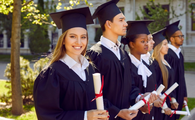 Nhóm sáu sinh viên tốt nghiệp -> đứng hàng dọc ngoài trời -> mặc áo choàng (gown) đen, mũ tốt nghiệp (mortarboard) vuông, cầm bằng cuộn trắng buộc ruy băng đỏ -> trước tòa nhà trường, ánh nắng ban ngà