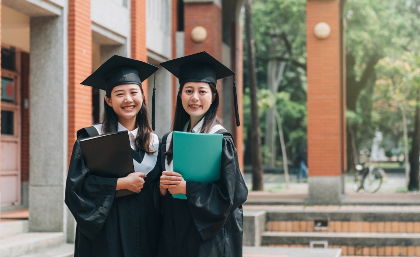 Hai nữ sinh viên mặc áo tốt nghiệp đen và mũ phớt, tay cầm bằng tốt nghiệp màu đen và xanh lá, đứng trước tòa nhà gạch đỏ với ánh sáng tự nhiên.