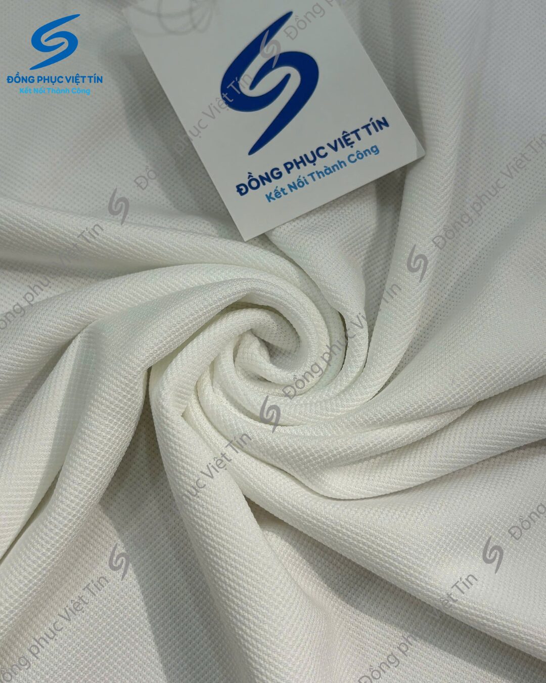 Vải thun màu trắng có bề mặt dệt lỗ kim, được xếp nếp xoắn ốc ở trung tâm. Phía trên có thẻ bài in logo Đồng Phục Việt Tín, hình ảnh có watermark thương hiệu lặp lại.