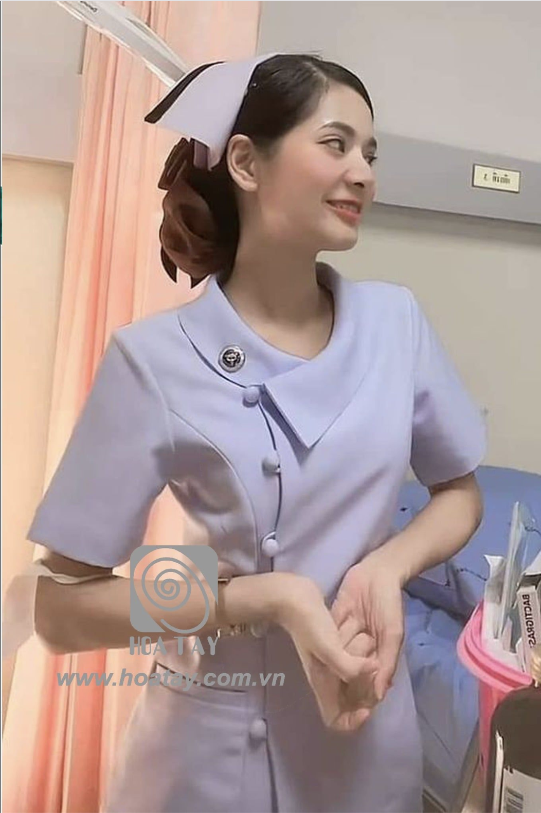 Một nữ điều dưỡng đang đứng, mặc áo đồng phục màu tím nhạt, cổ bẻ lệch (cổ chéo), cài khuy bọc vải, đội mũ y tế trắng, bối tóc gọn gàng, bối cảnh phòng bệnh viện với rèm hồng, ánh sáng trong nhà.