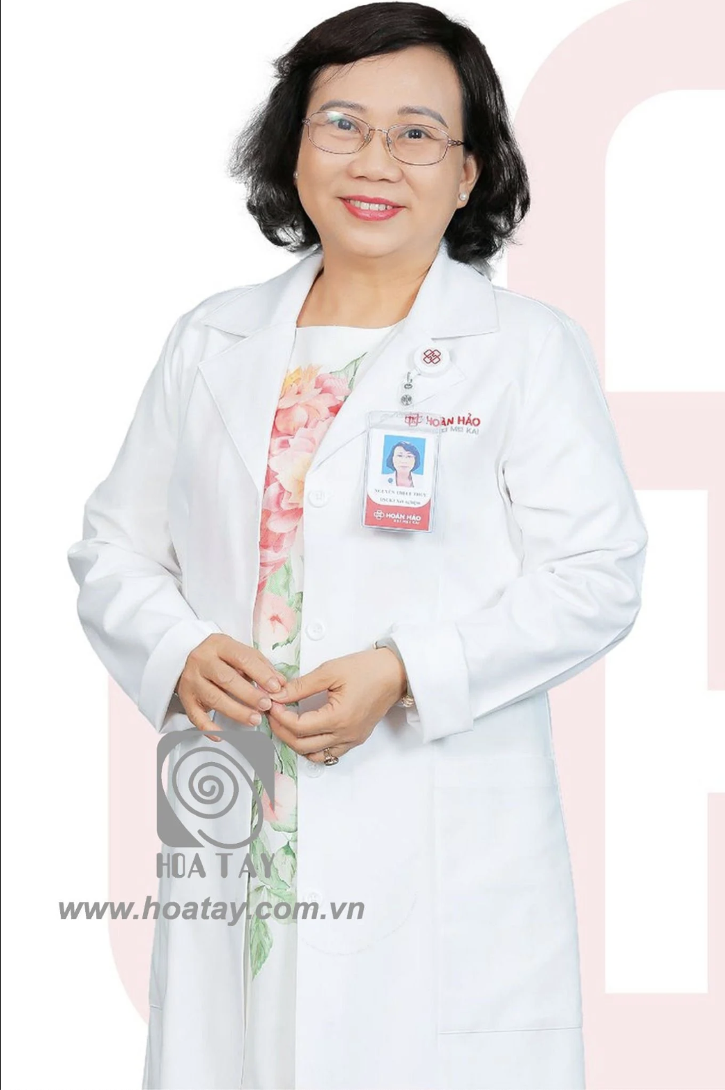 Một nữ bác sĩ đứng mỉm cười, mặc áo blouse trắng (áo khoác y tế) cổ bẻ, chất vải cotton dày dặn, đeo thẻ tên có logo bệnh viện, bên trong là váy hoa, phông nền studio sáng trắng.