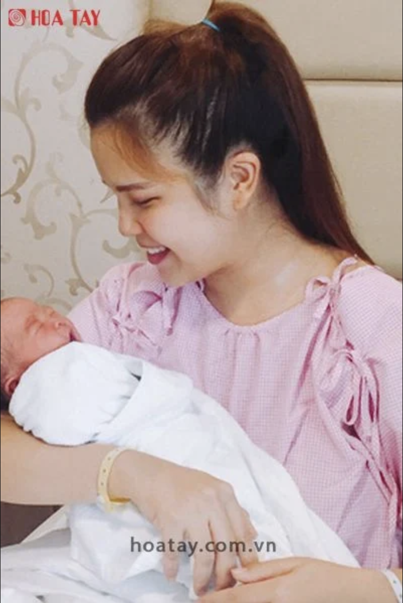 Một người mẹ và một trẻ sơ sinh (em bé) -> đang âu yếm, bế bồng -> mẹ mặc áo blouse hồng vải xô (muslin) thắt nơ vai, bé quấn khăn trắng (swaddle) -> bối cảnh trong nhà, ánh sáng tự nhiên dịu nhẹ.