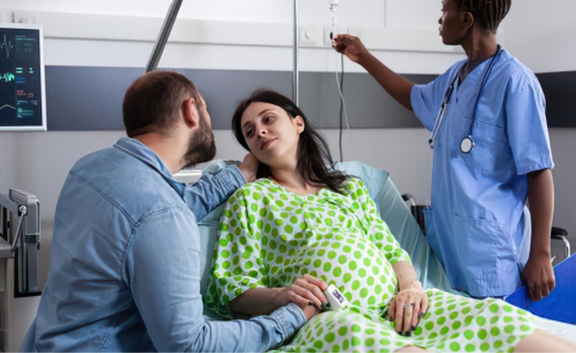 Bà bầu nằm trên giường bệnh, chồng ngồi bên cạnh, y tá đứng điều chỉnh truyền dịch. Bệnh nhân mặc áo bệnh nhân họa tiết chấm bi xanh lá, y tá mặc áo blouse xanh dương, có ống nghe. Bối cảnh phòng bệnh