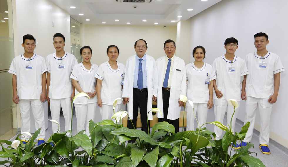 Nhóm 9 nhân viên y tế đứng thành hàng trong hành lang bệnh viện. Họ mặc đồng phục trắng với viền cổ xanh, áo blouse trắng, và hai người đàn ông lớn tuổi mặc áo choàng trắng (white coat) với cà vạt xan