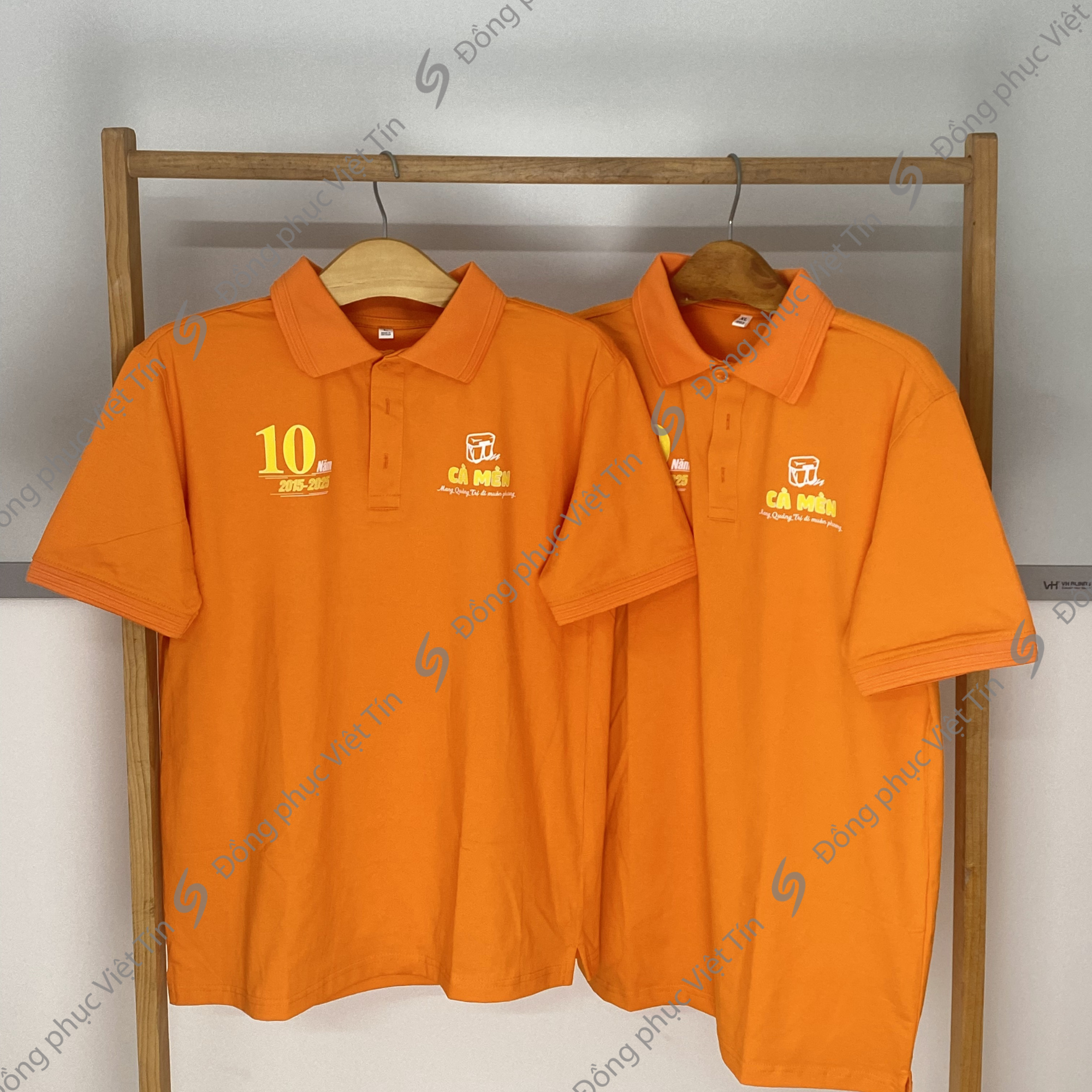 Hai chiếc áo polo màu cam dòng Fast-Fit, chất liệu Cotton 100%, tươi sáng treo trên giá gỗ. Áo có in logo 'CÀ MÈN' và kỷ niệm 10 năm (2015-2025) ở ngực trái. Bố cục cân đối, chuyên nghiệp.