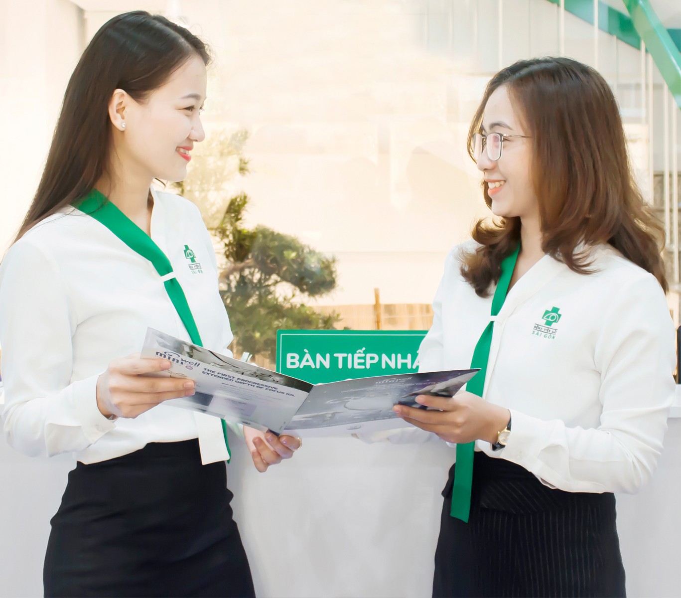 Hai nữ nhân viên y tế đang tư vấn, mặc áo sơ mi trắng cổ chữ V phối dải lụa xanh lá, chân váy đen, logo bệnh viện thêu trên ngực, đứng tại quầy lễ tân trong không gian sáng sủa.