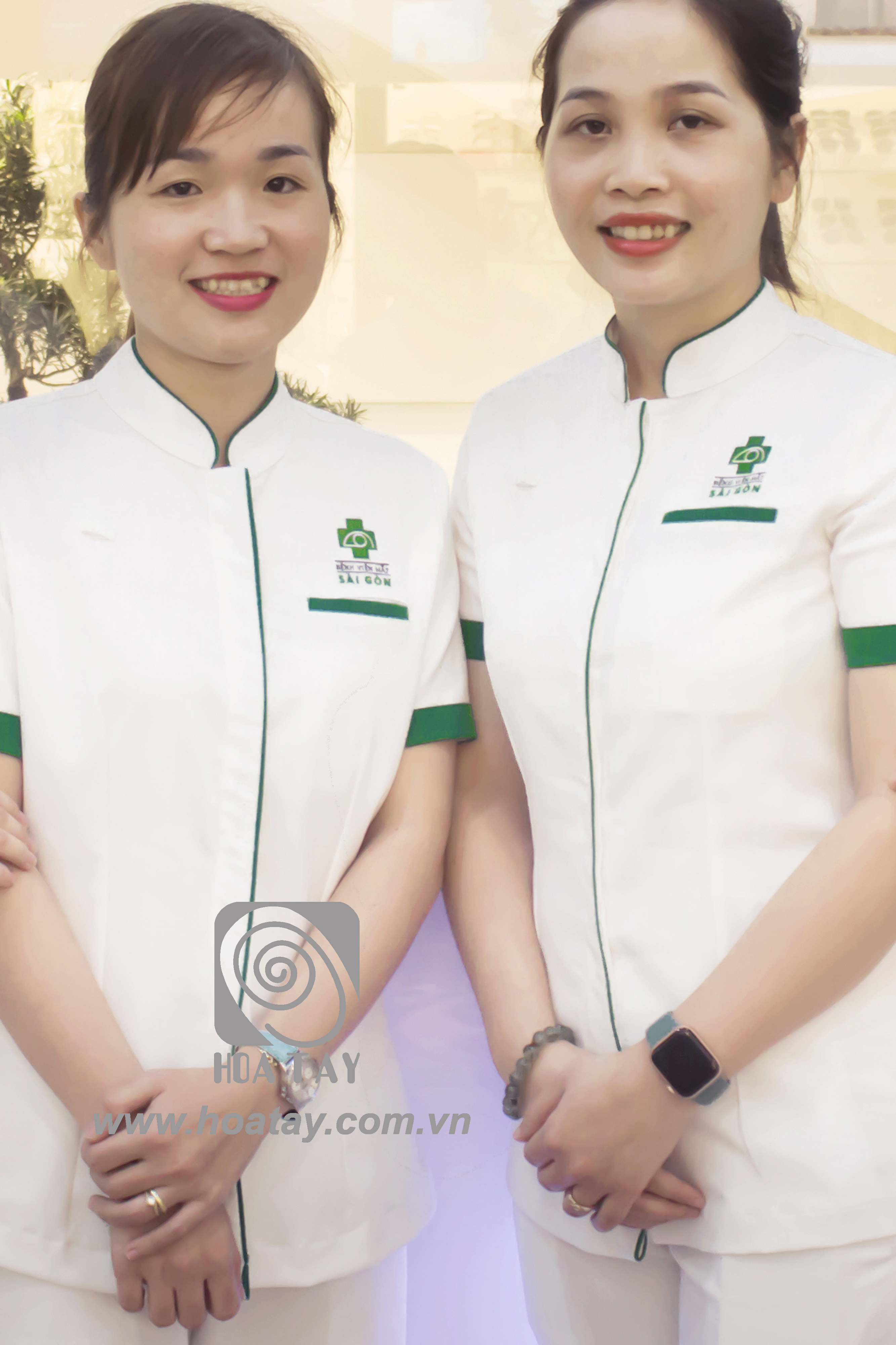 Hai nữ nhân viên y tế đang đứng mỉm cười; mặc áo blouse trắng cổ tàu (cổ đứng) viền xanh, tay ngắn, chất liệu vải phẳng phiu, logo bệnh viện thêu ngực trái; bối cảnh văn phòng sáng sủa.