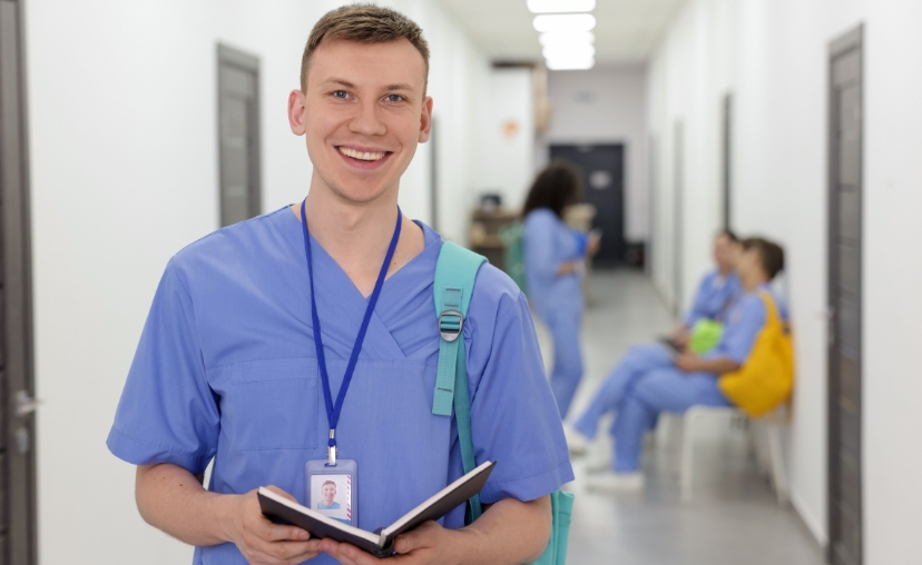 Một nam y tá trẻ mỉm cười, mặc bộ đồng phục (scrubs) màu xanh dương cổ chữ V, đeo thẻ nhân viên và ba lô xanh ngọc, cầm cuốn sổ tay, đứng trong hành lang bệnh viện sáng sủa.