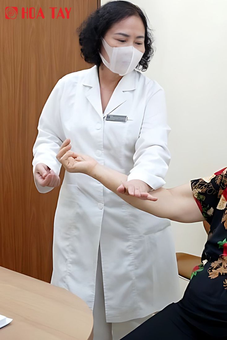 Một nữ bác sĩ đang khám tay cho bệnh nhân; mặc áo blouse trắng (áo khoác y tế) dài tay, cổ bẻ (cổ ve), cài khuy giữa, chất liệu vải cotton, đeo khẩu trang y tế; bối cảnh phòng khám, ánh sáng trong nhà