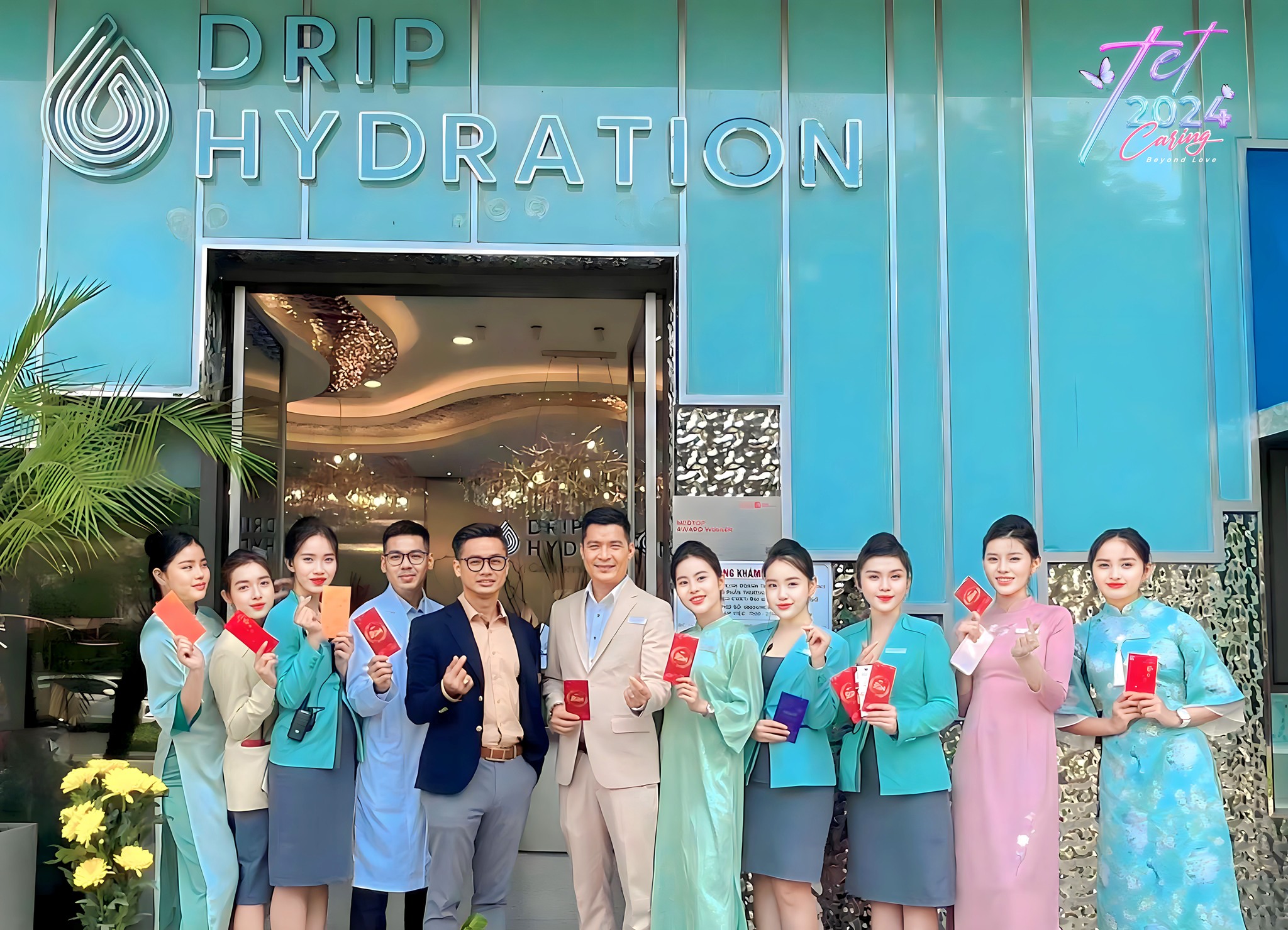 Nhóm 11 nhân viên đứng trước cửa hàng, cầm bao lì xì đỏ; mặc áo dài truyền thống, vest công sở, áo blouse trắng; bảng hiệu Drip Hydration màu xanh ngọc, ánh sáng ban ngày ngoài trời.