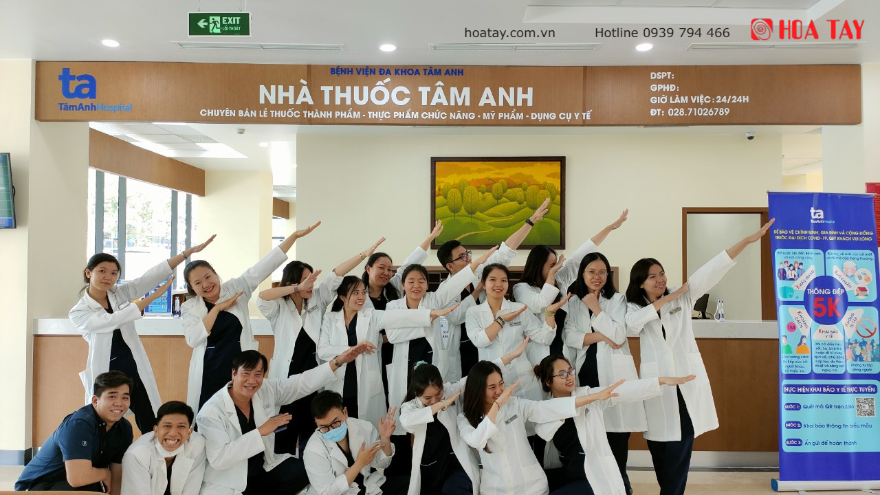 Nhóm khoảng 18 nhân viên y tế mặc áo blouse trắng, tạo dáng đồng bộ với tay duỗi ra. Bối cảnh là quầy lễ tân gỗ với biển hiệu 'NHÀ THUỐC TÂM ANH' và logo 'HÒA TAY'. Ánh sáng văn phòng sáng, có banner