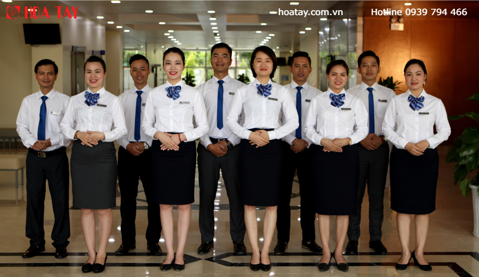Nhóm 9 nhân viên (5 nữ, 4 nam) đứng hàng ngang trong sảnh. Nữ mặc áo sơ mi trắng, váy đen, khăn lụa xanh caro; nam mặc áo trắng, cà vạt xanh, quần tây đen. Nền là sàn gạch bóng và logo Hoa Tay.