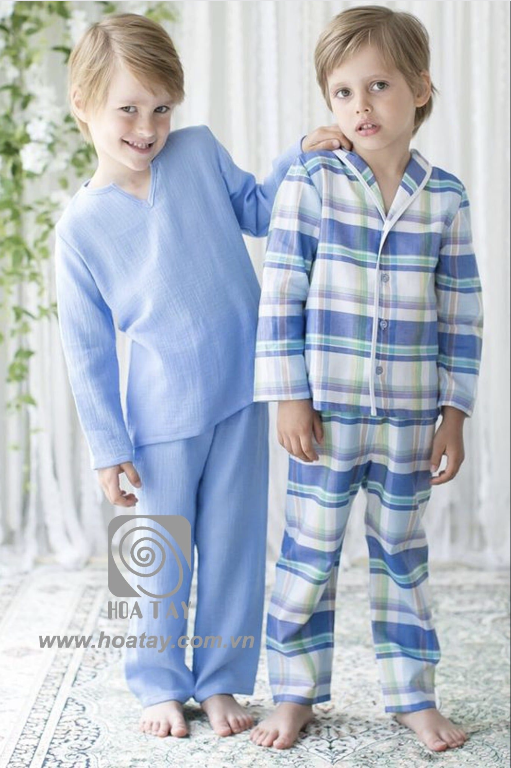 Hai bé trai đứng cạnh nhau, mặc đồ ngủ (pajamas) chất liệu vải xô (muslin) mềm mại, một bé mặc bộ trơn màu xanh dương cổ chữ V, một bé mặc bộ kẻ sọc (plaid) cài khuy, bối cảnh trong nhà ánh sáng tự nh
