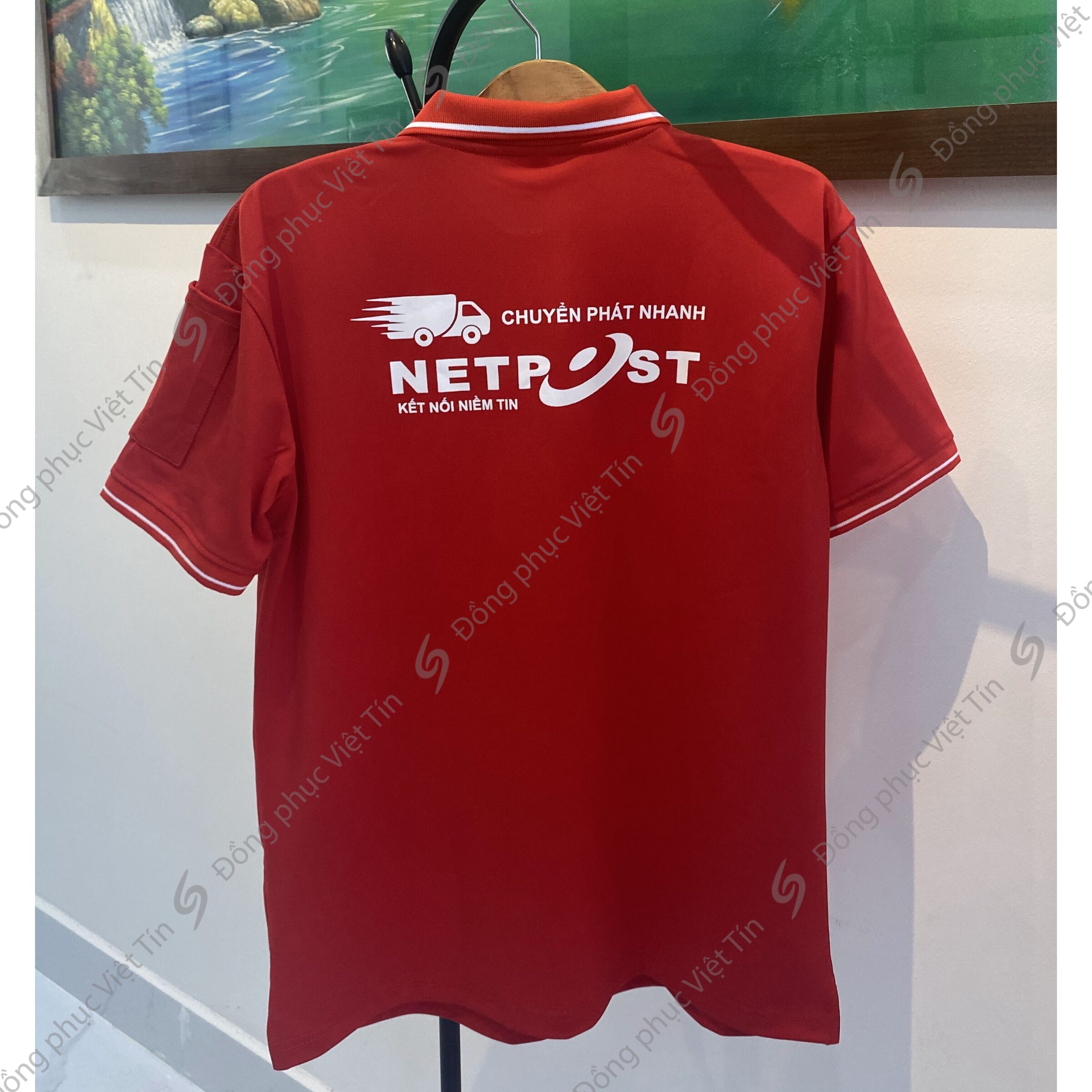 Áo polo màu đỏ tươi, mặt sau in logo trắng của thương hiệu Netpost với hình xe tải và dòng chữ "Chuyển phát nhanh". Áo được treo trên móc trong không gian nội thất.