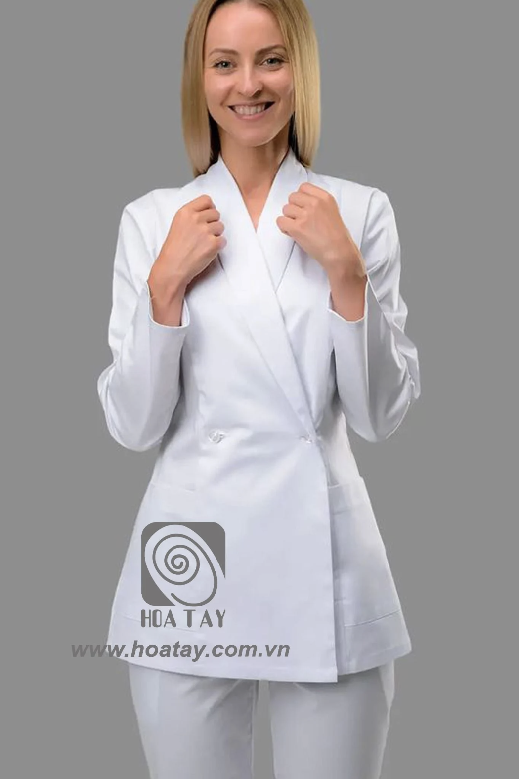 Một nữ nhân viên y tế đang đứng, mặc áo blouse trắng cổ vest (cổ bẻ) dáng dài, thiết kế cài khuy chéo (double-breasted), chất liệu vải trơn, tay dài, phông nền xám trung tính, ánh sáng studio đều.