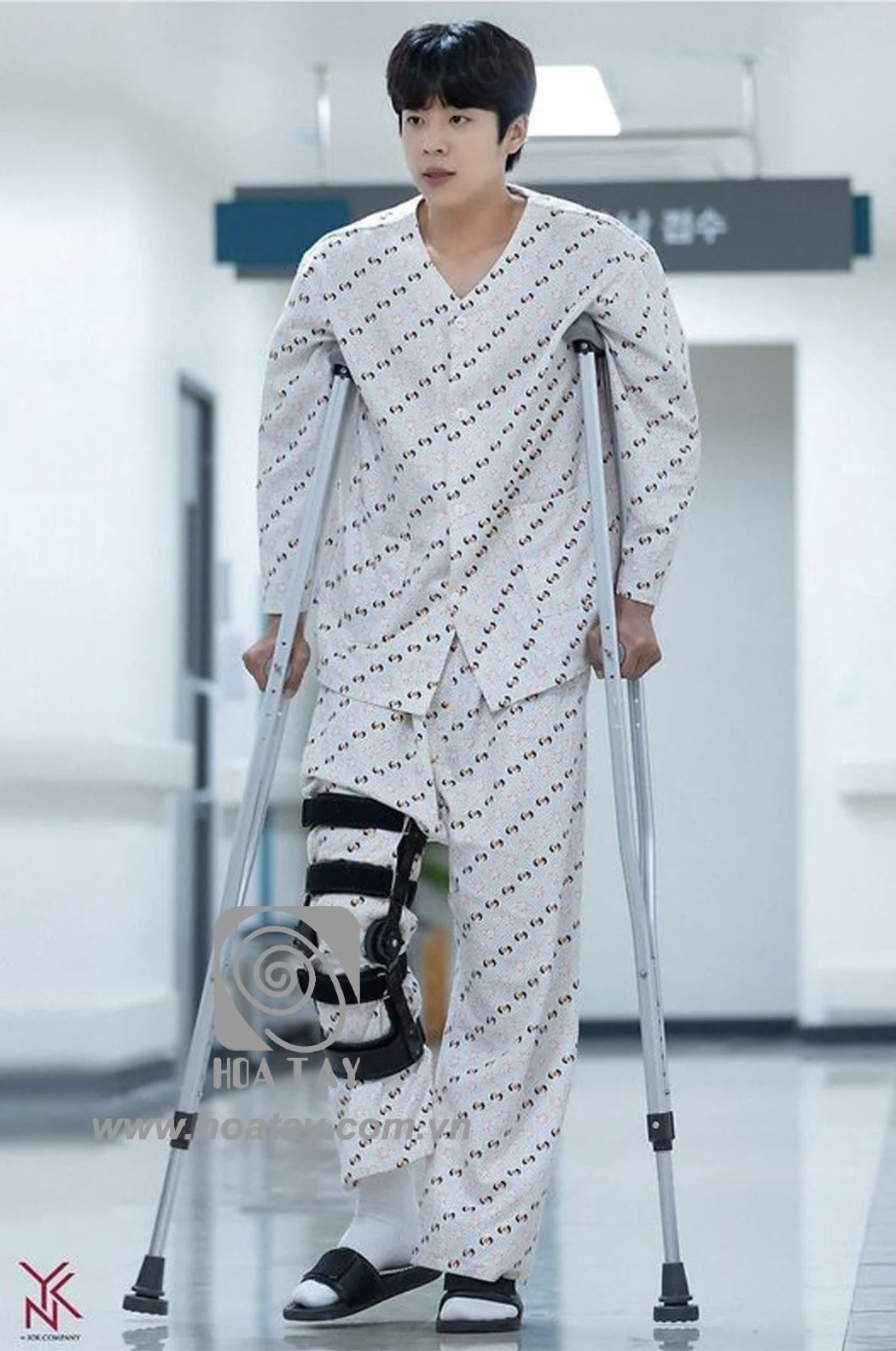 Một nam thanh niên đang đi bộ bằng nạng (gậy chống), mặc bộ đồ bệnh nhân (pyjama) họa tiết in, nẹp đầu gối (khung cố định) màu đen, dép quai ngang, trong hành lang bệnh viện với ánh sáng trắng.