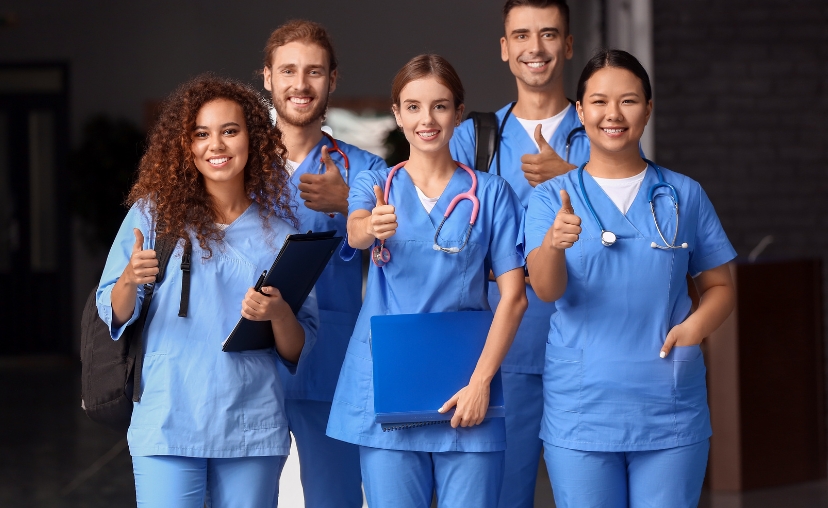 Năm nhân viên y tế (ba nữ, hai nam) đứng thành hàng, mỉm cười và giơ ngón cái, trong hành lang bệnh viện. Đồng phục y tá xanh dương, cổ chữ V, tay ngắn, quần dài. Hai người đeo ống nghe, một người cầm