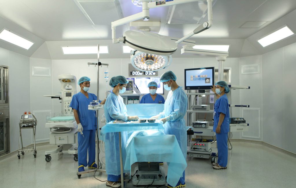 Năm nhân viên y tế trong phòng mổ -> đang phẫu thuật -> áo phẫu thuật xanh dương, mũ trùm đầu, khẩu trang, găng tay, đèn mổ trần tròn, màn hình theo dõi, bàn mổ phủ vải xanh -> phòng mổ trắng, ánh sán