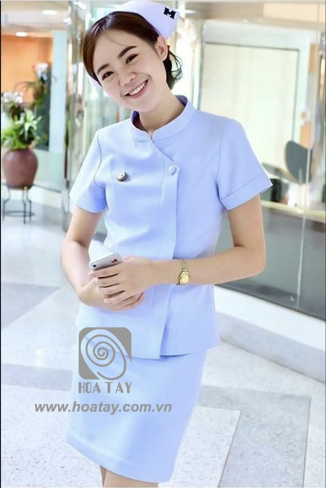 Một nữ y tá đang đứng, mặc đồng phục màu xanh nhạt (light blue), áo cổ tàu (mandarin collar) cài khuy lệch, tay ngắn, chân váy bút chì, đội mũ y tá, bối cảnh hành lang bệnh viện sáng sủa.
