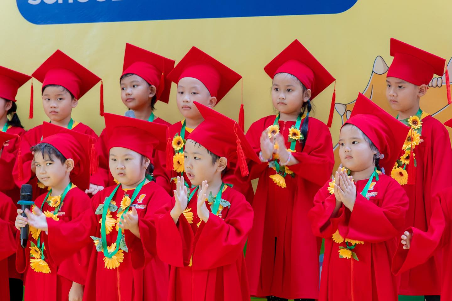 Nhóm 10 trẻ mầm non đang hát, mặc áo cử nhân (áo tốt nghiệp) đỏ rực, đội mũ vuông (mũ tốt nghiệp) có tua rua, đeo vòng hoa hướng dương vàng, đứng trên sân khấu dưới ánh sáng đèn vàng.