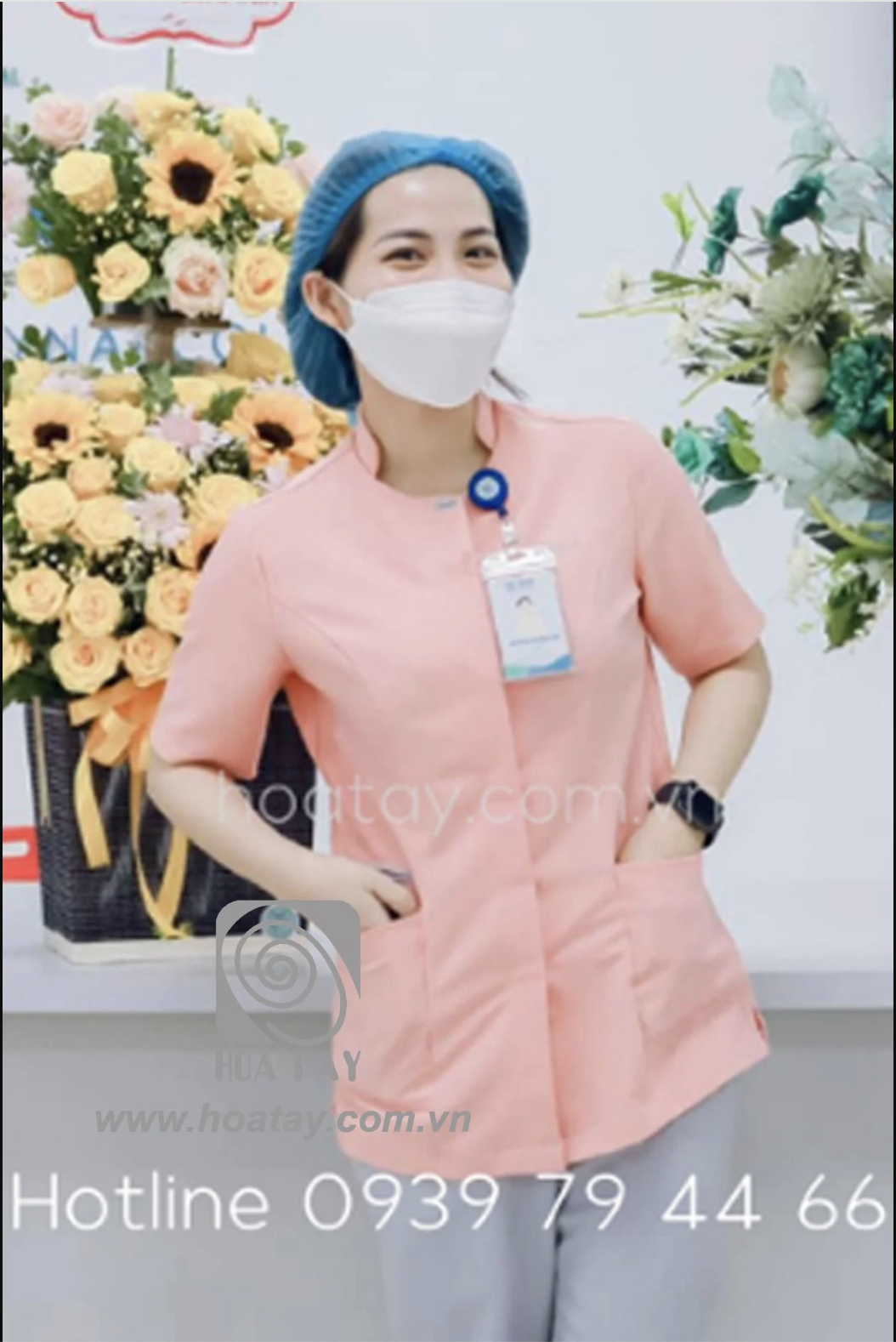 Một nữ nhân viên y tế đang đứng, mặc áo blouse cổ tròn (cổ tàu) màu hồng phấn, tay ngắn, chất liệu vải phẳng phiu, có hai túi lớn phía trước, đeo thẻ tên và mũ bao tóc y tế, bối cảnh văn phòng sáng sủ