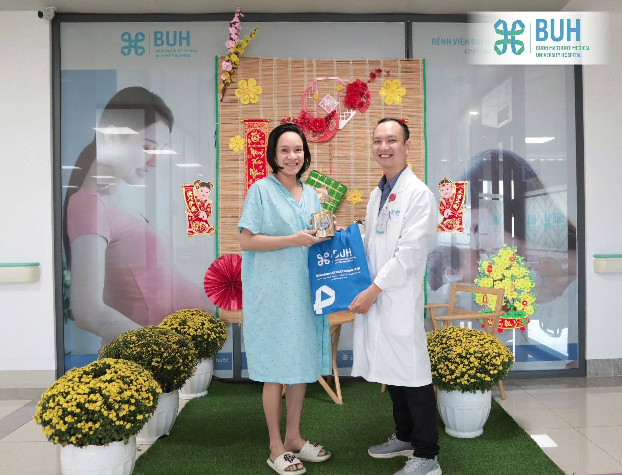 Một bác sĩ nam và một bệnh nhân nữ đang đứng; bác sĩ mặc áo blouse trắng (áo khoác y tế) dài tay, bệnh nhân mặc váy bệnh viện màu xanh nhạt, bối cảnh trang trí Tết với hoa cúc vàng và phông nền tre tr