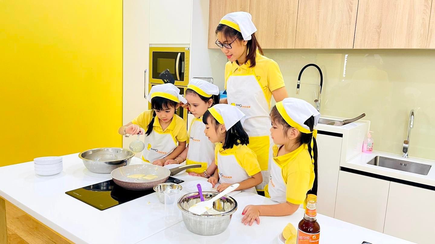 Một giáo viên và bốn trẻ em đang nấu ăn; mặc tạp dề (yếm bảo hộ) trắng viền vàng, mũ đầu bếp (nón vải) trắng, áo polo vàng; trong bếp hiện đại, ánh sáng tự nhiên.