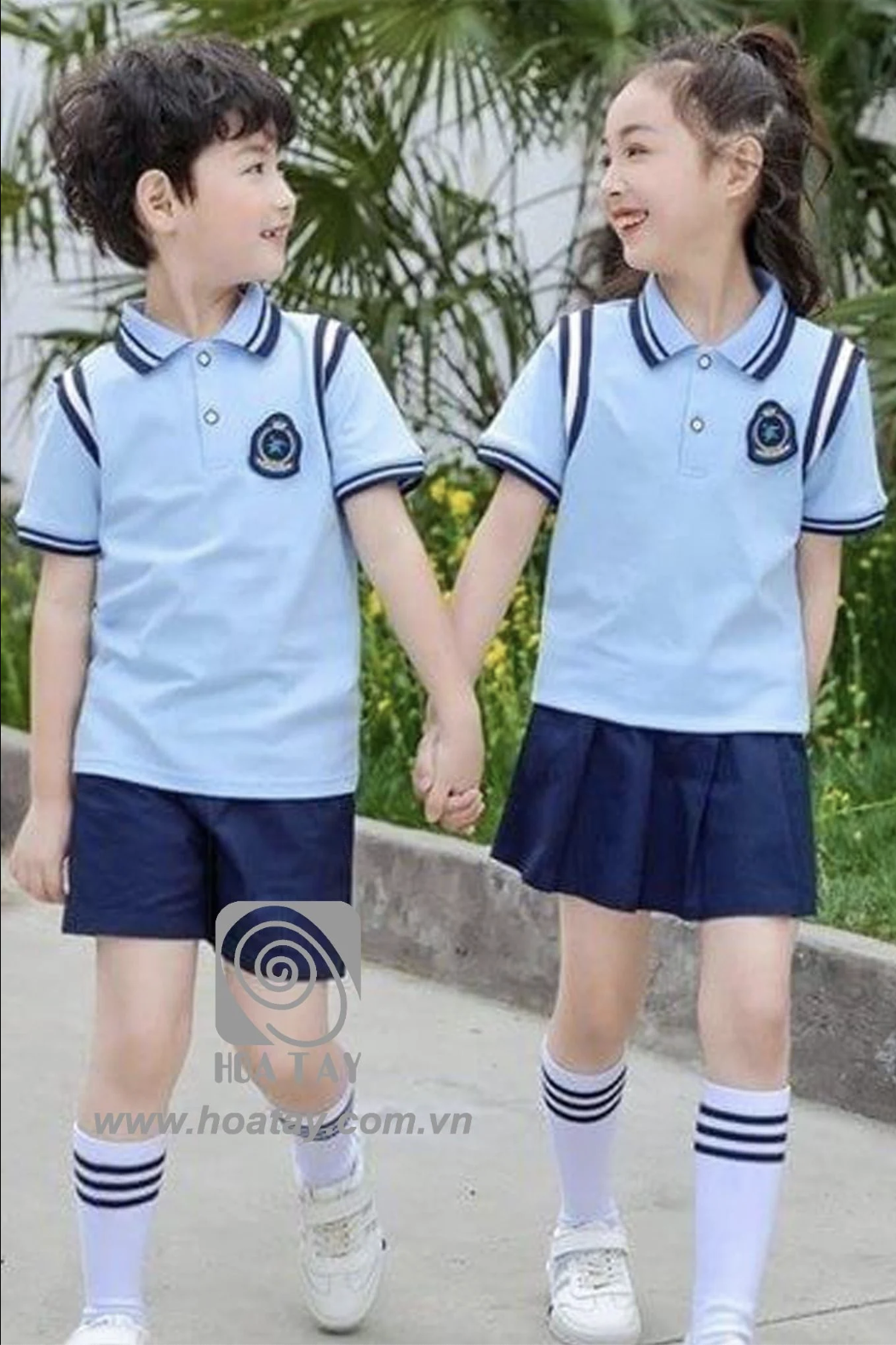 Hai học sinh (một nam, một nữ) đang nắm tay nhau; mặc áo polo xanh nhạt cổ bẻ (cổ áo), quần đùi/váy xếp ly xanh đậm, tất trắng kẻ sọc; chất liệu vải cotton, logo thêu ngực; bối cảnh ngoài trời, ánh sá