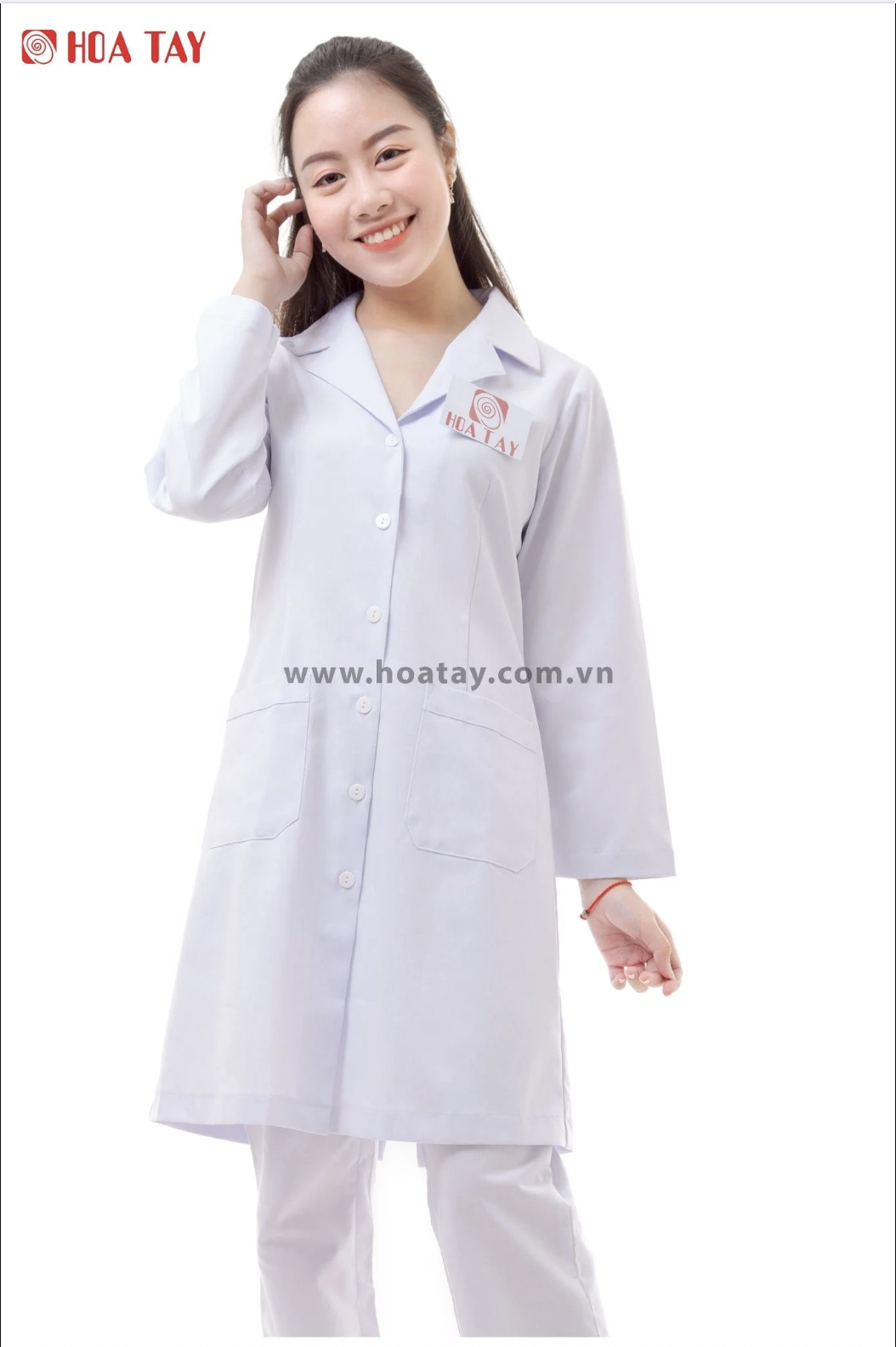 Một nữ nhân viên y tế đang đứng mỉm cười; mặc áo blouse trắng (áo khoác phòng thí nghiệm) dài tay, cổ bẻ (cổ ve), cài khuy giữa, có hai túi hông lớn; chất liệu vải phẳng, nền trắng sáng, ánh sáng stud