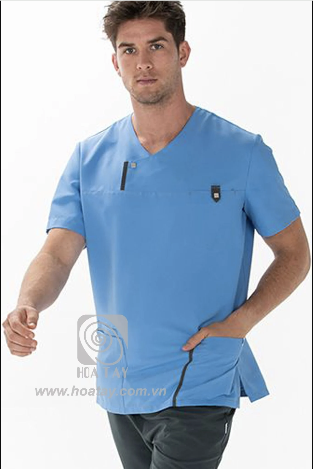 Một nam nhân viên y tế đang đứng. Áo blouse cổ chữ V (cổ tim), màu xanh dương, chất liệu vải trơn, có túi ngực phối khóa kéo và chi tiết viền đen tương phản. Phông nền trắng, ánh sáng studio đều.