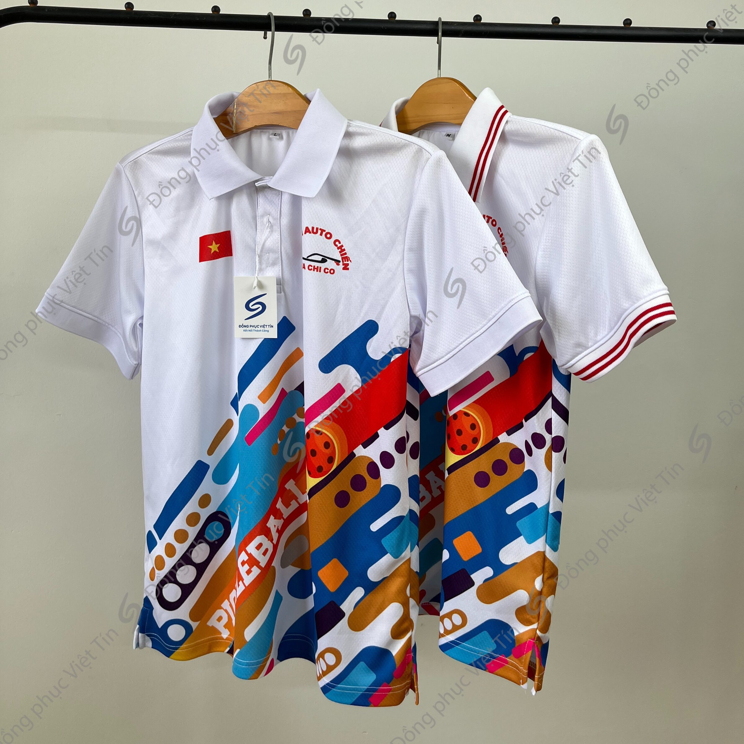 Hai chiếc áo polo trắng treo trên móc, họa tiết in màu sắc rực rỡ (đỏ, xanh, vàng) ở phần dưới, có logo Auto Chiến và cờ Việt Nam, chất liệu vải thể thao.