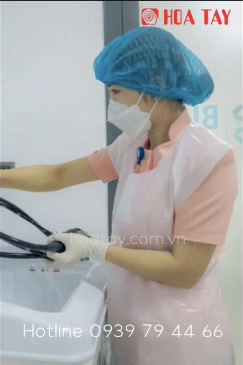 Một nhân viên y tế đang vệ sinh thiết bị nội soi, đội mũ trùm đầu (mũ y tế) màu xanh, đeo khẩu trang trắng, mặc áo blouse hồng và tạp dề nhựa (tấm chắn bảo vệ) trong phòng khám với ánh sáng trắng.