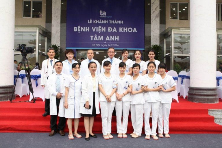 Nhóm khoảng 15 nhân viên y tế đứng trên thảm đỏ trước biển hiệu 'Lễ Khánh Thành Bệnh Viện Đa Khoa Tâm Anh'. Họ mặc áo blouse trắng, áo dài trắng và áo choàng y khoa, đứng thành hai hàng.