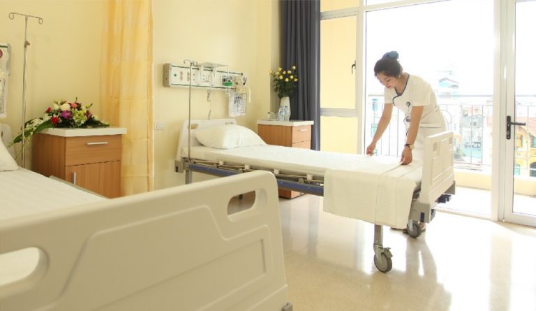 Y tá trong đồng phục trắng đang trải ga trên giường bệnh viện có bánh xe, khung kim loại, thành giường nhựa trắng, trong phòng bệnh có cửa kính lớn và ánh sáng tự nhiên.