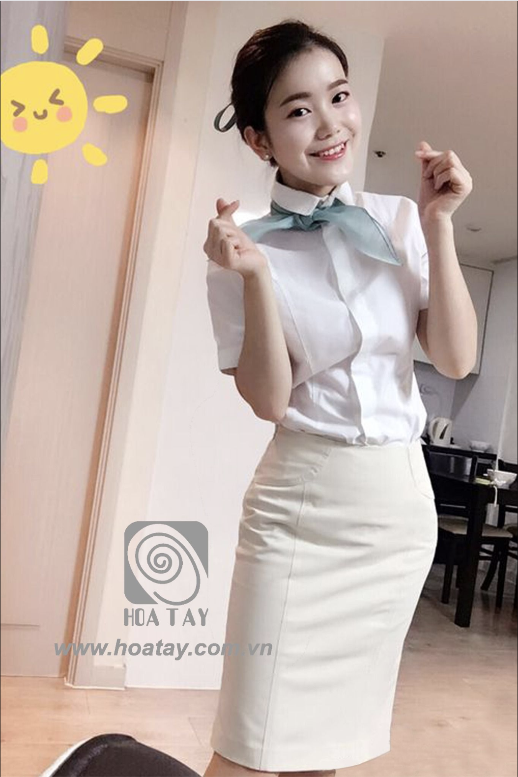 Một nữ nhân viên đang tạo dáng tay hình trái tim, mặc áo sơ mi trắng tay ngắn (cổ bẻ) phối cùng khăn quàng cổ màu xanh nhạt và chân váy bút chì (ôm sát) màu kem, bối cảnh văn phòng trong nhà với ánh sáng tự nhiên.