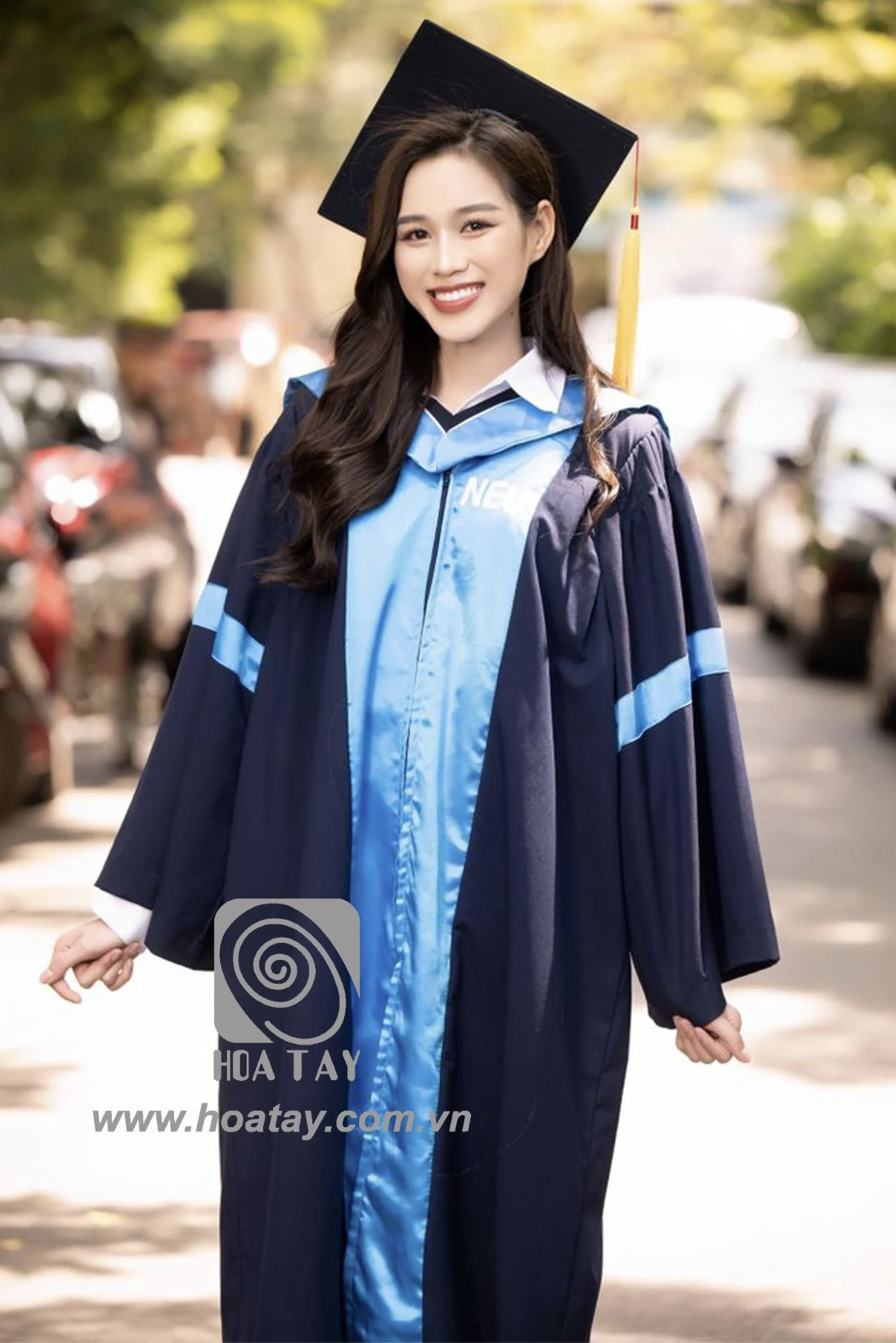Một nữ sinh viên đội mũ cử nhân (mũ tốt nghiệp vuông) đang mỉm cười, mặc áo lễ phục (áo choàng tốt nghiệp) màu xanh navy phối dải lụa xanh dương sáng, tay áo rộng, cổ áo sơ mi trắng bên trong, bối cản