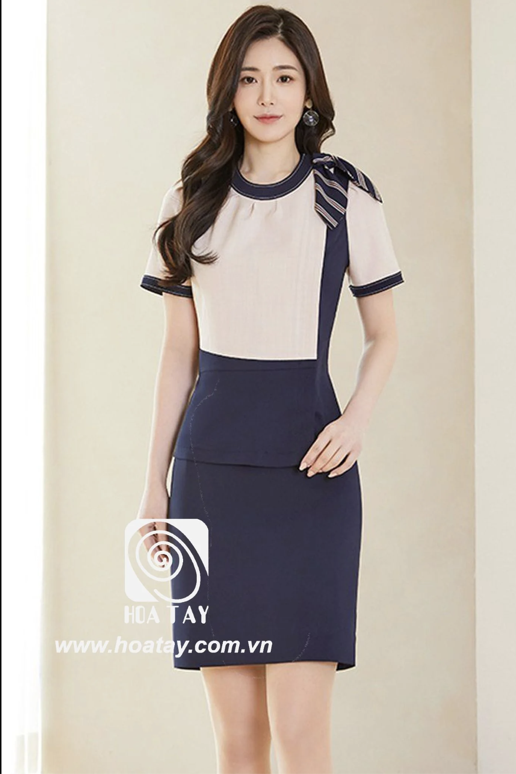 Một nữ nhân viên đứng tạo dáng, mặc bộ đồng phục phối màu (color-block) gồm áo cổ tròn tay ngắn màu kem và chân váy bút chì (váy ôm) màu xanh navy, điểm nhấn nơ kẻ sọc trên vai, chất liệu vải đứng for